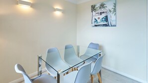 A modern dining area with a glass table and light-colored chairs, a painting of a van on the wall, and recessed lighting.