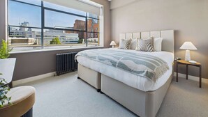 A modern bedroom with a large window, a bed with a striped blanket, two bedside tables with lamps, and a radiator. - Spacious and Bright Apartment by Tech City (Greater London)