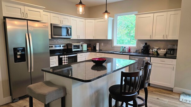 A modern kitchen with white cabinets, stainless steel appliances, a central island with bar stools, and a large window providing natural light.