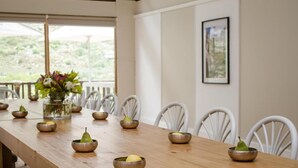 Une longue table à manger en bois, sur laquelle sont disposés plusieurs bols remplis de poires et de citrons, des chaises blanches, un vase avec des fleurs et un tableau encadré accroché au mur.