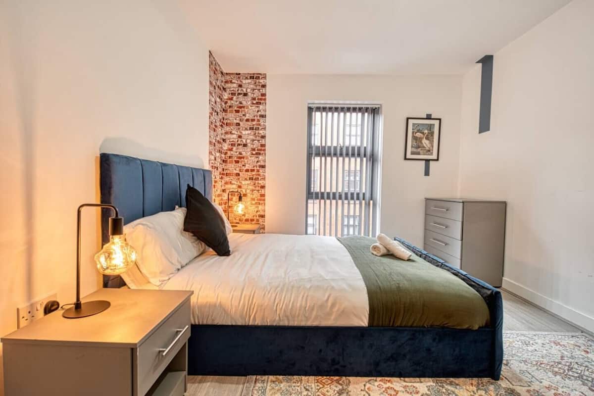 Une chambre à coucher moderne avec un mur d’accent en briques, un grand lit avec une tête de lit bleue, une lampe de chevet et une commode.