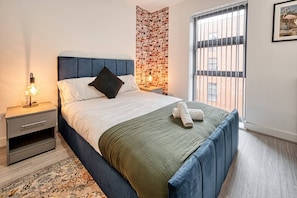 A modern bedroom with a large bed, a nightstand, a patterned rug, a brick accent wall, and a window with blinds. - Jewellery Courts (Birmingham)