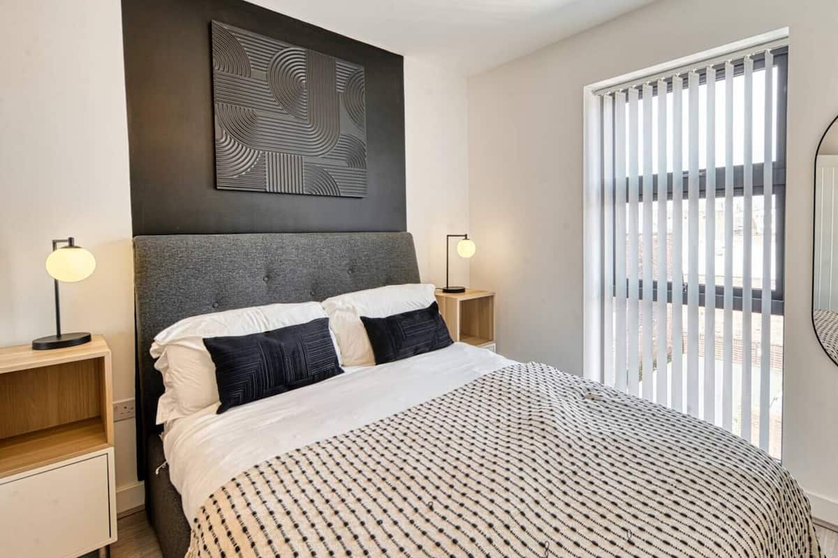 Une chambre moderne avec un grand lit, deux tables de chevet, une œuvre d’art accrochée au mur et une fenêtre équipée de stores verticaux.