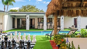 Zona de piscina con piezas de ajedrez, mobiliario exterior y una estructura cubierta de paja.