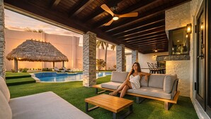 Zona junto a la piscina con una sombrilla de paja, un sofá y una mesa de madera.