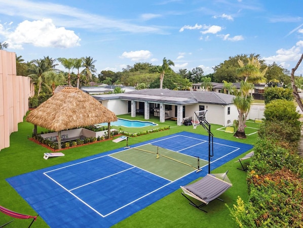 A tennis court with a net, a pool, and a thatched-roof structure.