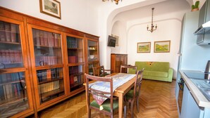 Una biblioteca con estanterías de madera, una mesa de comedor con sillas, un sofá verde y una zona de cocina.