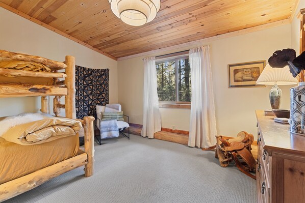A rustic room with a bunk bed, a chair, a window with curtains, a lamp, and a framed picture on the wall.