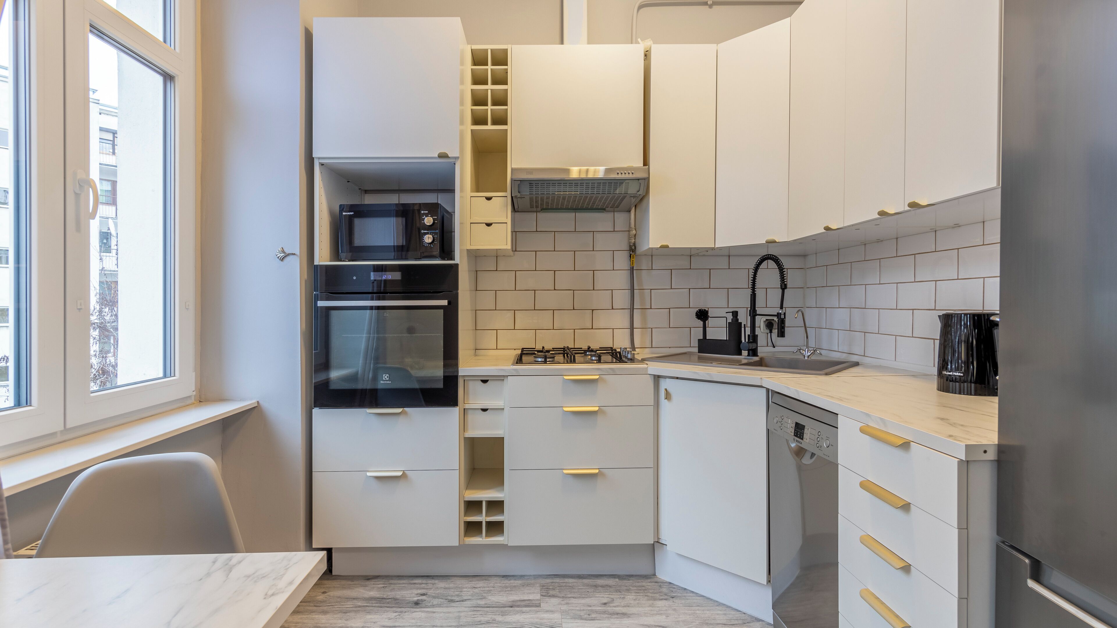 Una cocina compacta con muebles blancos, horno empotrado, microondas, estufa a gas y pileta con grifo moderno.