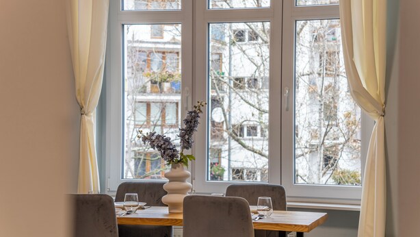 A dining area with a table set for four, a vase with flowers, and a view of buildings through large windows. - Apartamenty Sun & Snow Szczytnicka (Wroclaw)