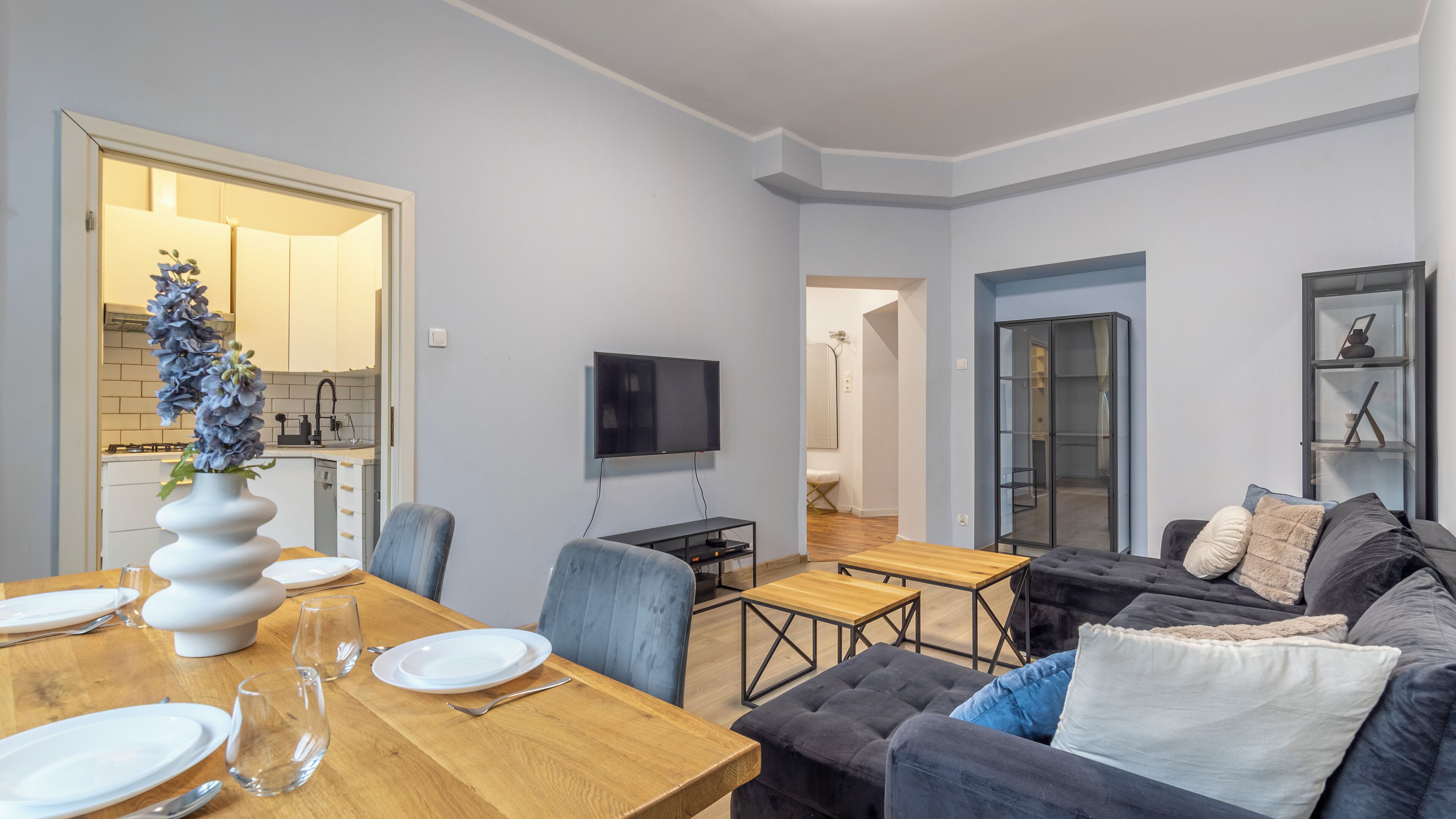 A modern living room with a dining area, a flat-screen TV, and a kitchen visible through an open door.