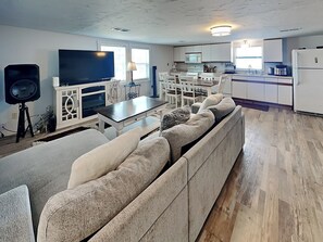 A modern living room with a large flat-screen TV, a sofa, a dining table, and a kitchen area with white cabinets and appliances. - Surfside Beach Bungalow | Water Views + Community Pool (Surfside Beach)
