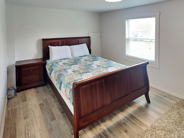 A bedroom with a wooden bed, a nightstand, a dresser, and a window with a view of buildings. - Surfside Beach Bungalow | Water Views + Community Pool (Surfside Beach)