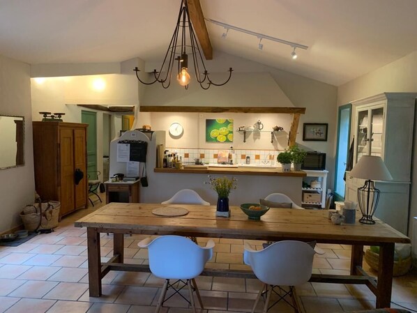 Uma área de refeições com mesa de madeira e cadeiras brancas, uma cozinha com relógio e obras de arte, e um lustre.
