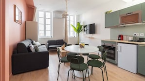 A modern living room with a sofa, a dining table with chairs, a kitchen area with a fridge, oven, and a TV mounted on the wall.