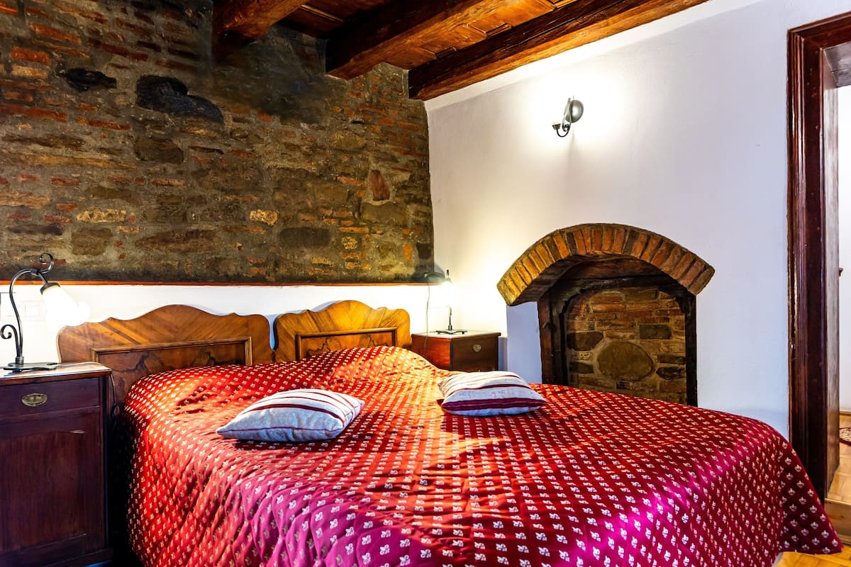 Une chambre avec une cheminée en briques, une tête de lit en bois et une couvre-lit à motifs rouges.