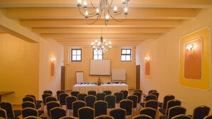 Un salón de conferencias con filas de sillas mirando una larga mesa, una araña y dos pantallas de proyección.
