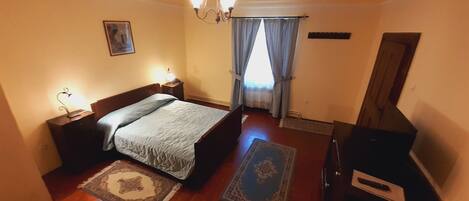 Un dormitorio con cama, mesitas de noche, cómoda, candelabro y ventana con cortinas.