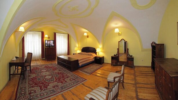 Un dormitorio con una cama grande, un escritorio, una silla, un aparador y un espejo. La habitación tiene piso de madera y una alfombra estampada.