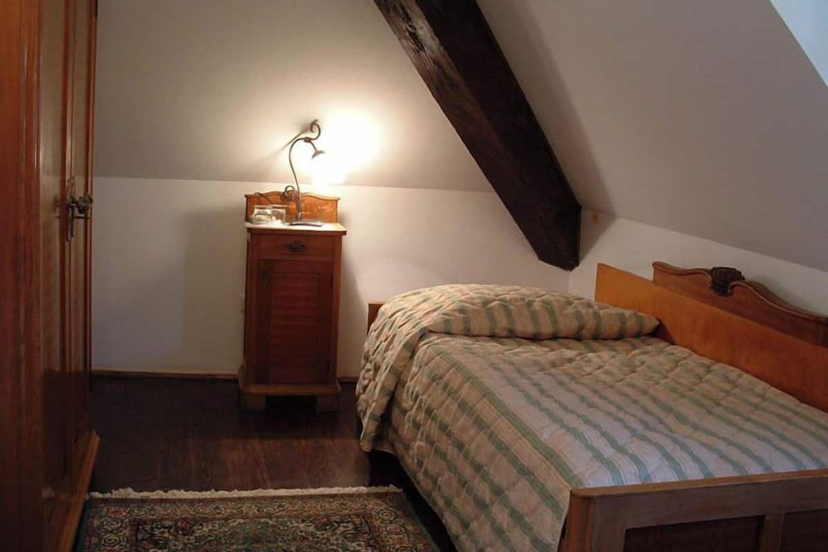 Une petite chambre à coucher accueillante, avec un plafond en pente, une table de chevet en bois surmontée d’une lampe, un lit avec une literie à rayures et un tapis à motifs.