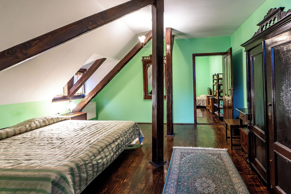 Une chambre avec un plafond en pente, un lit recouvert d’une couvre-lit à motifs, une armoire en bois et un miroir.