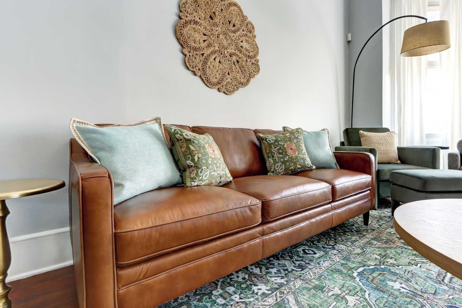A brown leather sofa with patterned cushions, a woven wall decoration, and a patterned rug in a well-lit room with a floor lamp.