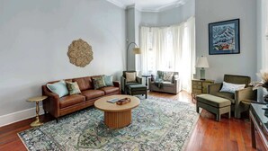 A living room with a brown leather sofa, a round coffee table, a patterned rug, a green armchair, a side table, and a painting on the wall.