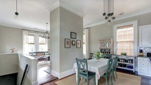 A dining area with a table set for four, a kitchen with white cabinets, and a living room with a sofa and a window. - The Koi Escape, Victorian | Upper Porch | Parking (Savannah)
