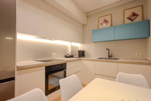 Una cucina moderna con mobili bianchi, forno integrato, microonde e lavello. C'è un tavolo da pranzo con sedie e un armadietto blu sopra il lavello.