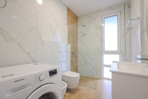 A modern bathroom with a washing machine, toilet, glass shower enclosure, and a large window. - Sanpietro Vacation Apartment 047 (Ishëm)