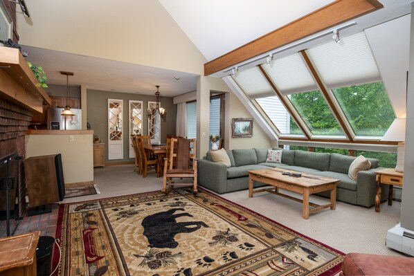 A spacious living area with a high ceiling, a dining area, and a kitchen. There are large windows providing natural light and a view of the outdoors.