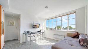 A modern living room with a large window, a flat-screen TV, a dining area with chairs, and a sofa. - Roland House - Deluxe Studio (London)
