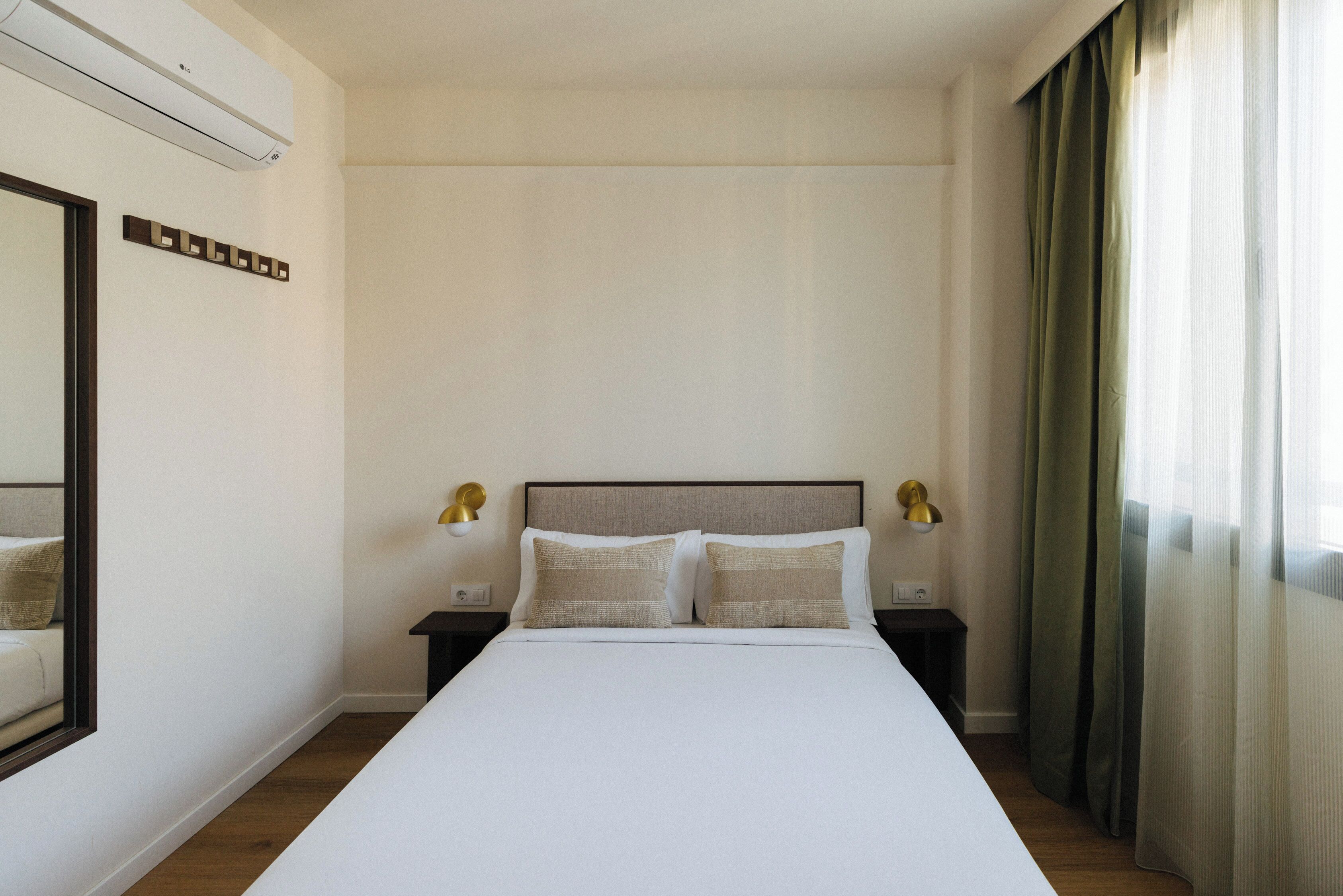 Una habitación de hotel con una cama grande, dos mesitas de noche con lámparas, un espejo y un ventanal con cortinas.