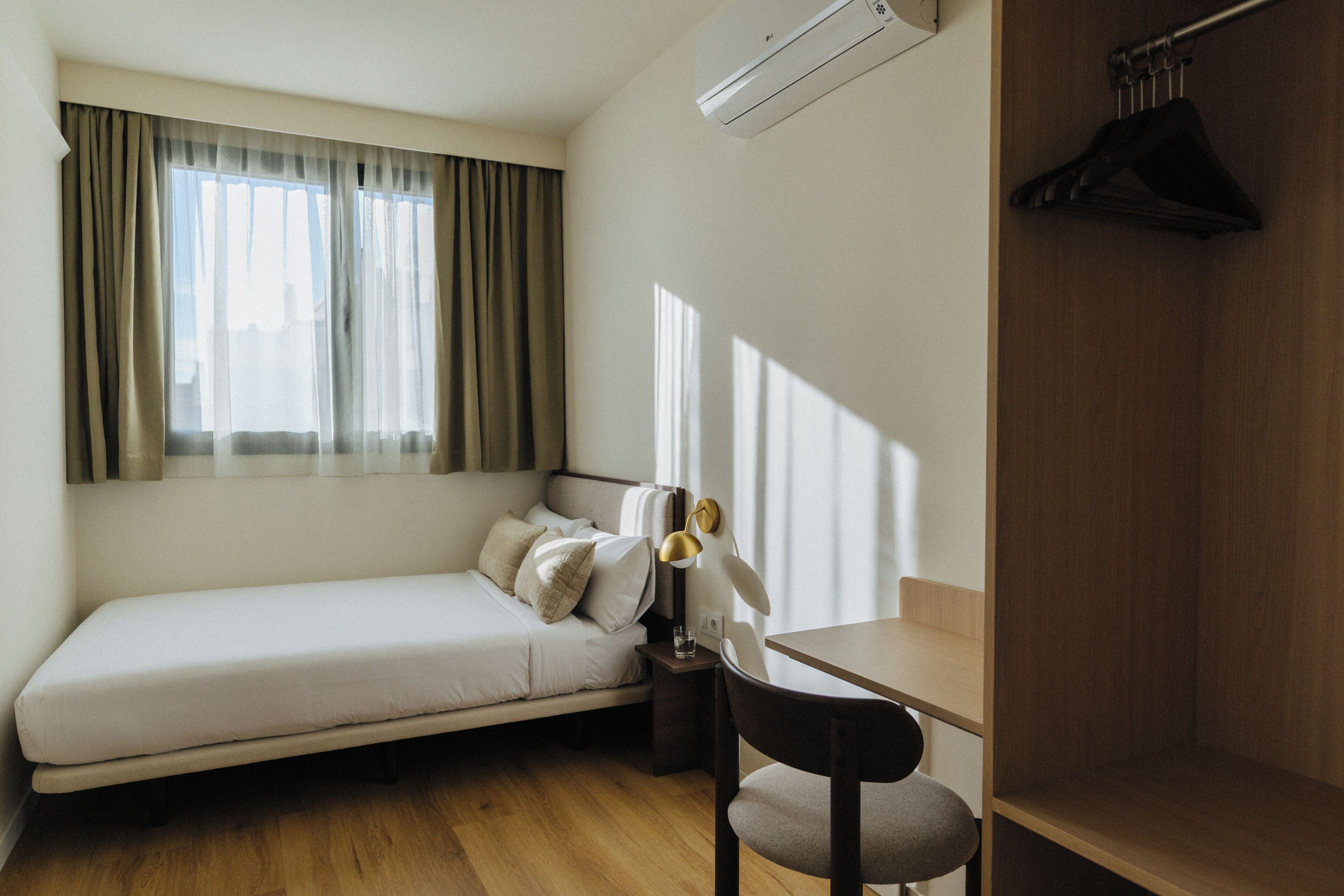 A compact room with a single bed, a small table, a chair, and a hanging coat rack.