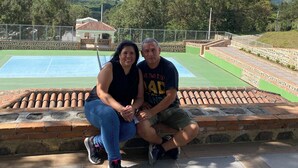 Dos personas sentadas en un muro de ladrillo mirando una cancha de tenis con montañas al fondo.