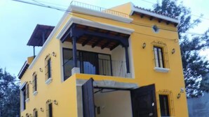 Un edificio amarillo con balcón, puerta negra y un pequeño jardín.