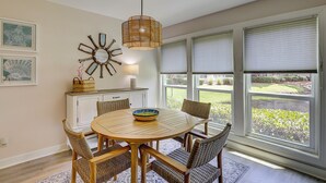 A dining area with a round wooden table, wicker chairs, a hanging light fixture, and a view through large windows. - Sunset Rentals | Lighthouse Tennis 2387 (Hilton Head Island)
