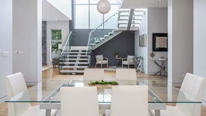 En modern inredning med ett glasbord i matsalen, vita stolar, en trappa och en hängande lampa.