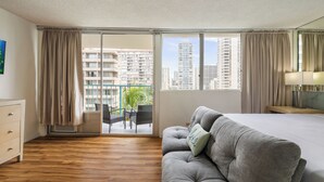 A hotel room with a bed, a sofa, a desk, a chair, and a view of tall buildings through large windows.