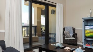 Un salon avec une porte-fenêtre coulissante donnant sur un balcon, un fauteuil en osier, un téléviseur et une petite table avec des télécommandes.