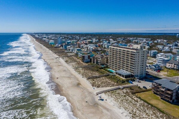 An aerial view of a coastal town with a beachfront hotel featuring multiple floors, a parking area, and a boardwalk leading to the beach.