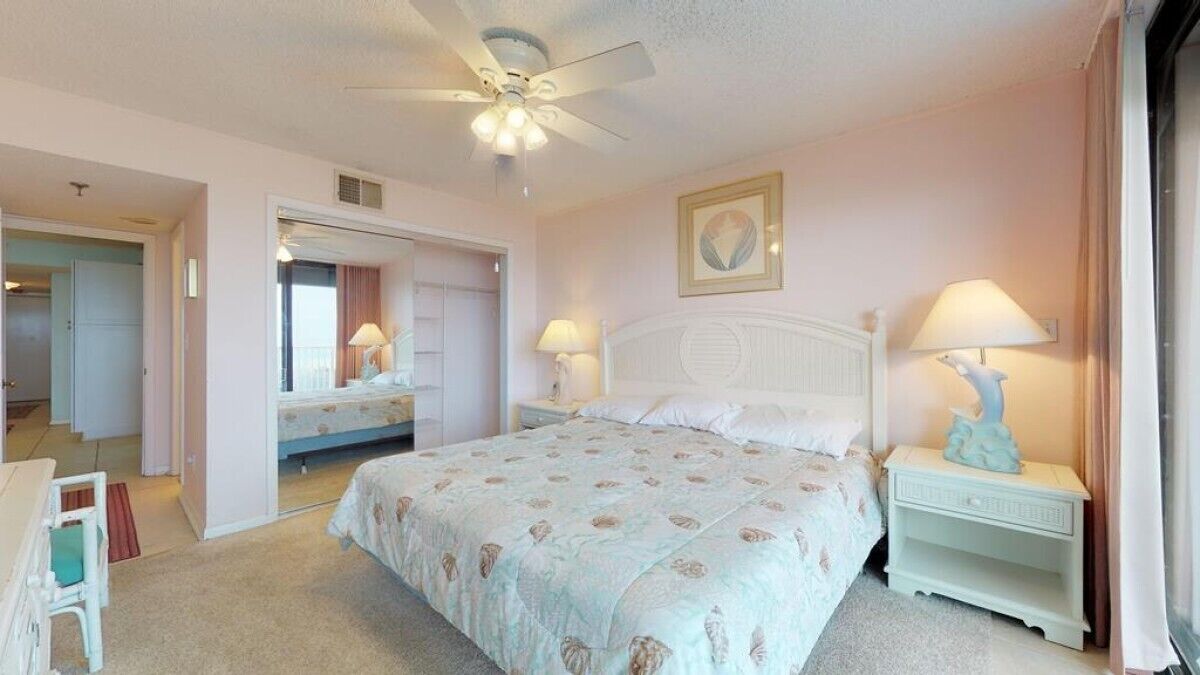 A bedroom with a large bed, two bedside tables with lamps, a ceiling fan, and a door leading to another room.