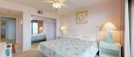 A bedroom with a large bed, two bedside tables with lamps, a ceiling fan, and a door leading to another room.