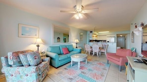 A living room with a blue sofa, a pink armchair, and a patterned armchair. There is a dining area with a table and chairs, and a kitchen in the background.