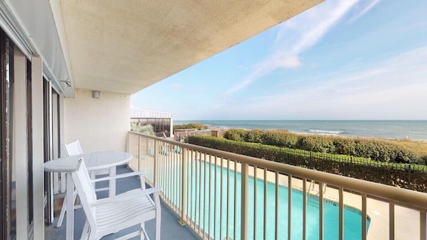 A balcony with a pool, white chairs, and a view of the ocean.