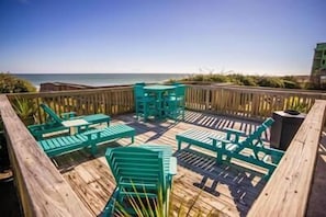 A wooden deck with green outdoor furniture, including chairs and a table, overlooking the ocean.