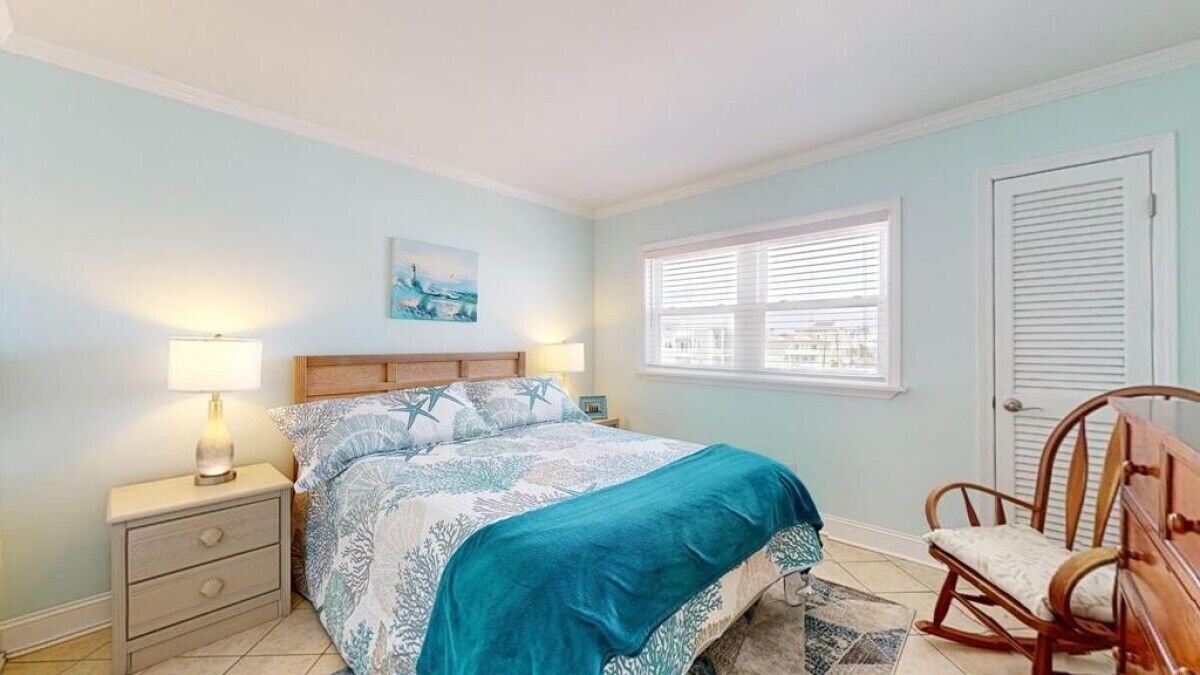 Une chambre à coucher avec un lit, deux lampes de chevet, un fauteuil à bascule, une fenêtre avec des stores et une porte.