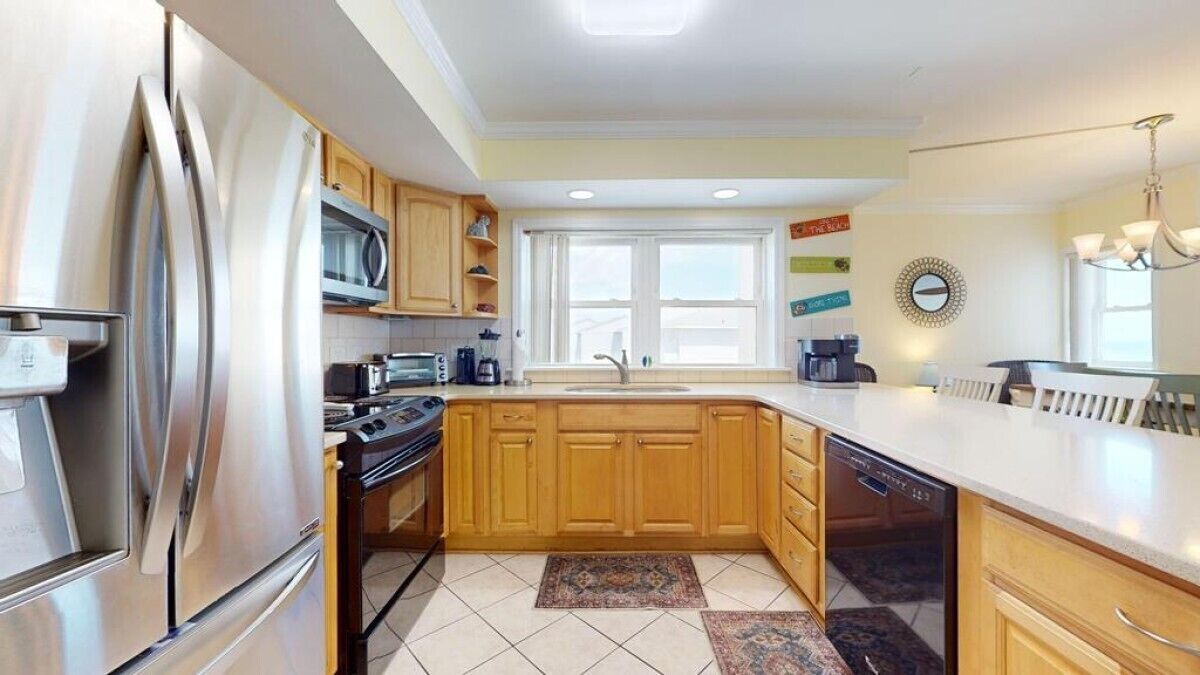 A modern kitchen with stainless steel appliances, wooden cabinets, a large window, and a chandelier.
