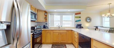 A modern kitchen with stainless steel appliances, wooden cabinets, a large window, and a chandelier.
