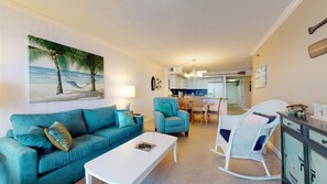 Una sala de estar con un sofá azul aguamarina, una mesa de centro blanca y un sillón mecedor. En la pared, hay un cuadro con una escena de playa.